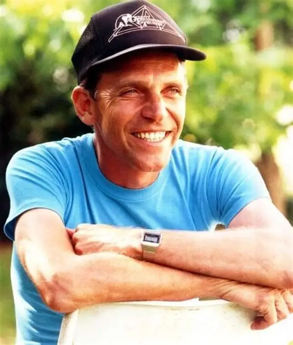 A man with a wide smile and a black baseball cap is leaning forward, resting his arms on a white chair. He is wearing a bright blue t-shirt and a digital watch. The background shows lush green foliage. This image is of director Sam Firstenberg, who was interviewed on OH!CAST.