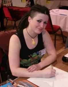 Tanya Roberts drawing at a comic convention table, wearing a Teenage Mutant Ninja Turtles tank top and guest badge.